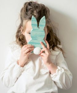 Cute little girl in white blouse covering face with Easter rabbit toy while standing near white wall